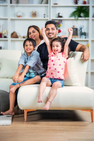 Téléchargez les photos : Portrait d'une jeune famille indienne heureuse de quatre personnes assise sur un canapé dans le salon - en image libre de droit