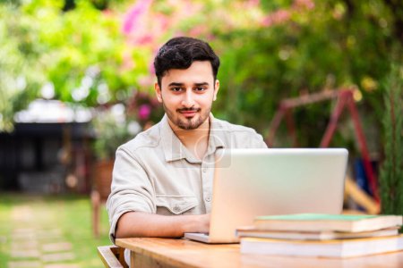Téléchargez les photos : Asiatique indien mâle étudiant ou pigiste en utilisant ordinateur portable tout en étant assis sur la table à l'extérieur - en image libre de droit