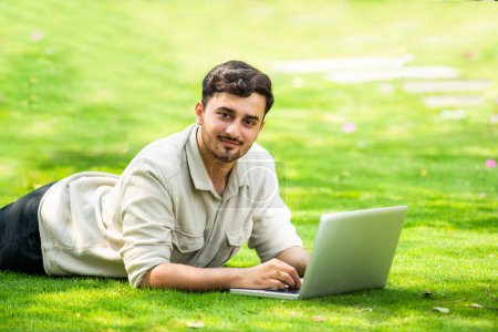 Téléchargez les photos : Jeune beau indien asiatique l'homme à l'aide d'ordinateur portable dans le parc tout en étant couché sur l'herbe - en image libre de droit