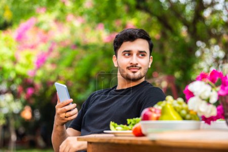 Photo for Indian young man sitting and using smartphone outdoors - Royalty Free Image