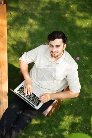 Overhead view of handsome indian man using laptop in the garden at table looking up into camera
