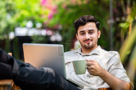 Photo for Asian Indian male student or freelancer using laptop computer while sitting on table outdoors - Royalty Free Image
