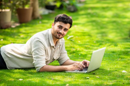 Photo for Young handsome Indian asian man using laptop in the park while lying on the grass - Royalty Free Image
