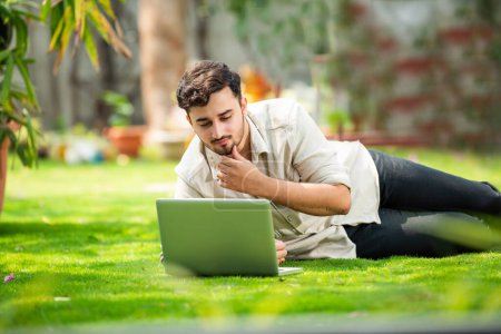 Foto de Joven guapo indio asiático hombre utilizando portátil en el parque mientras que la mentira en la hierba - Imagen libre de derechos