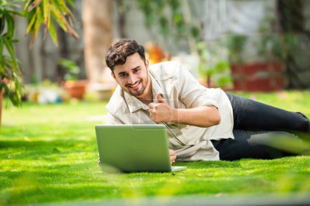 Téléchargez les photos : Jeune beau indien asiatique l'homme à l'aide d'ordinateur portable dans le parc tout en étant couché sur l'herbe - en image libre de droit