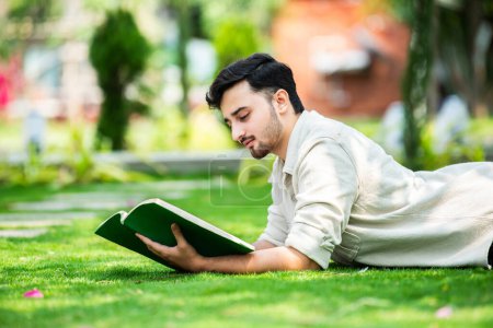 Foto de Joven estudiante masculino indio atento se encuentra en la hierba verde y lee libro en el parque verde de verano - Imagen libre de derechos