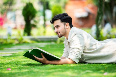 Téléchargez les photos : Jeune étudiant indien attentif se trouve sur l'herbe verte et lit le livre au parc vert d'été - en image libre de droit