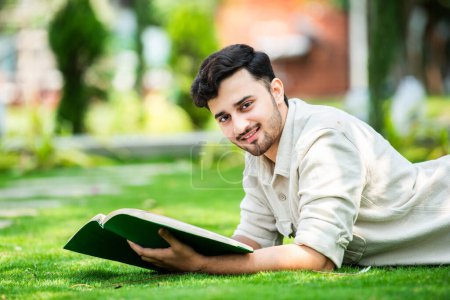 Foto de Joven estudiante masculino indio atento se encuentra en la hierba verde y lee libro en el parque verde de verano - Imagen libre de derechos
