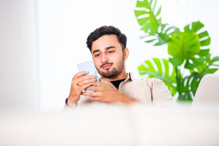 Photo for Young Indian handsome man using smartphone in living room while sitting on sofa - Royalty Free Image