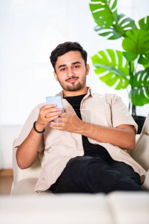 Téléchargez les photos : Jeune homme beau indien utilisant un smartphone dans le salon tout en étant assis sur le canapé - en image libre de droit