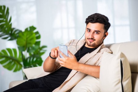 Photo for Young Indian handsome man using smartphone in living room while sitting on sofa - Royalty Free Image