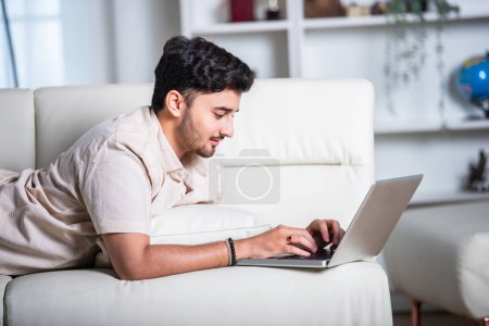 Photo for Indian man working on laptop at home while lying on sofa - Royalty Free Image