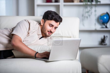 Téléchargez les photos : Indien travaillant sur ordinateur portable à la maison tout en étant couché sur le canapé - en image libre de droit