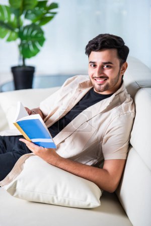 Foto de Indio asiático guapo joven lee libro en casa en sofá - Imagen libre de derechos