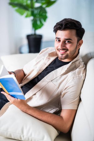 Foto de Indio asiático guapo joven lee libro en casa en sofá - Imagen libre de derechos