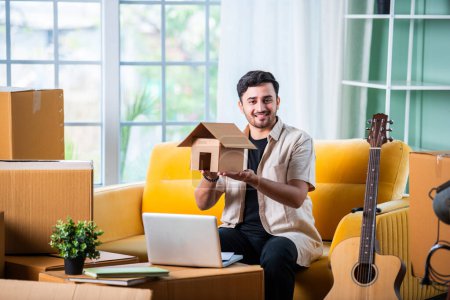 Photo for Indian man sitting on sofa with cardboard boxes, searching rental property on laptop or smartphone - Royalty Free Image