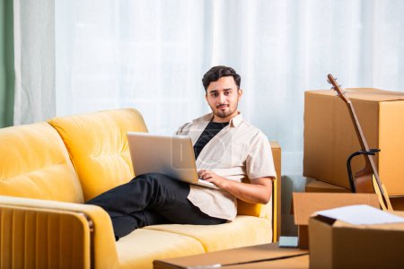 Indian man sitting on sofa with cardboard boxes, searching rental property on laptop or smartphone