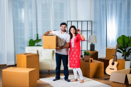 Photo for House Ownership. Young Indian Couple Showing Keys And Holding Cardboard Box in new home - Royalty Free Image