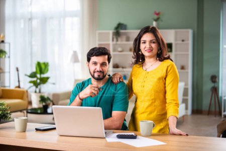 Téléchargez les photos : Heureux jeune couple marié indien assis sur la table calculer les dépenses utiliser un service bancaire en ligne facile à la maison - en image libre de droit