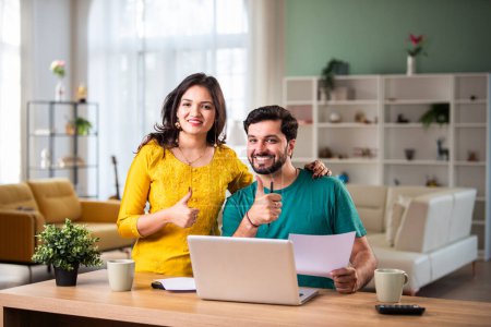 Téléchargez les photos : Heureux jeune couple marié indien assis sur la table calculer les dépenses utiliser un service bancaire en ligne facile à la maison - en image libre de droit