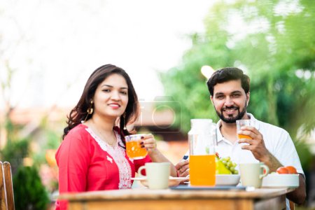 Photo for Indian husband and wife having fresh juice in breakfast while sitting outdoors at table - Royalty Free Image