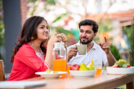 Téléchargez les photos : Jeune couple indien assis à la table du petit déjeuner à l'extérieur et prenant du thé ou du café chaud - en image libre de droit