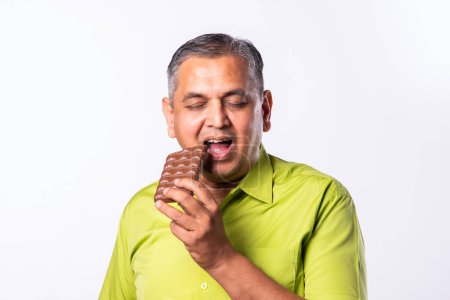 Foto de Asiático indio hombre en 50s disfrutando mientras comer chocolate en blanco fondo - Imagen libre de derechos