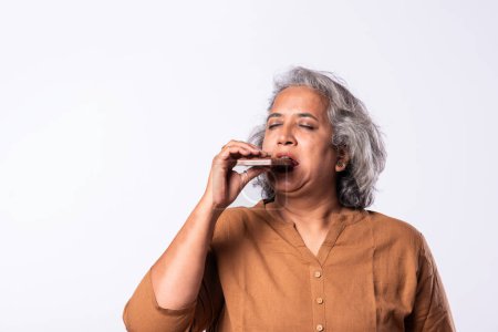 Foto de Asiática india mujer en 50s disfrutando mientras comer chocolate en blanco fondo - Imagen libre de derechos