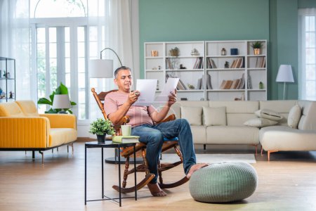 2. Home office. Confident Indian asian man checking financial reports while sitting on rocking chair in living room