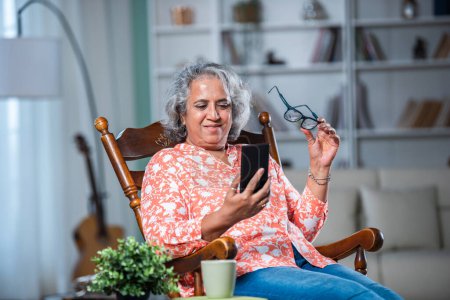 Téléchargez les photos : 1. Indien asiatique femme dans les années 40 avec des cheveux gris en utilisant smartphone à la maison, assis sur rocking fauteuil relaxant - en image libre de droit
