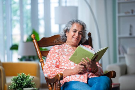 Téléchargez les photos : Indien asiatique moyen âge gris cheveux femme lecture un livre tandis que assis sur rocking chair dans salon - en image libre de droit