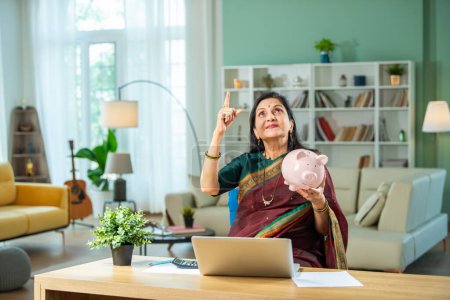 Photo for Indian asian old lady with piggy bank, laptop, money and 3d house model at home - Royalty Free Image