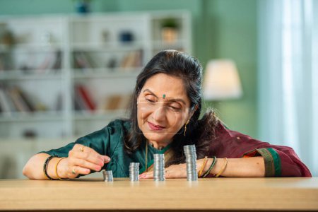 happy asian Indian senior married woman counting money tower on table