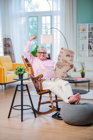 Téléchargez les photos : Indien asiatique vieil homme lecture nouvelles papier tandis que assis sur rocking chair à la maison - en image libre de droit