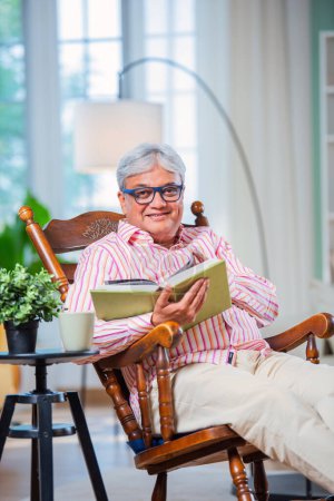 Téléchargez les photos : Indien asiatique vieil homme lecture livre ou roman tandis que assis sur rocking chair à la maison - en image libre de droit