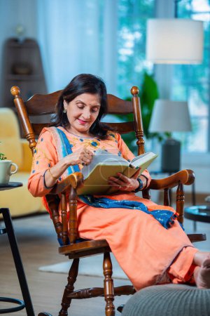 Téléchargez les photos : Indien asiatique retraité senior femme lecture un livre ou journal tandis que assis sur rocking chair dans salon - en image libre de droit