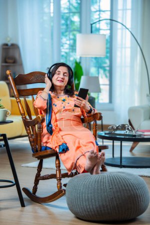 Téléchargez les photos : Heureux indien asiatique moyen âge femme en utilisant téléphone mobile tout en écoutant de la musique sur écouteurs à la maison - en image libre de droit