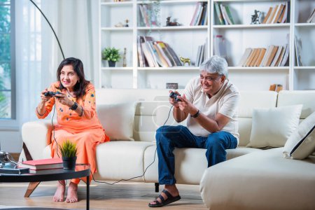 Photo for Indian Senior couple having fun playing video games while sitting on sofa in home - Royalty Free Image