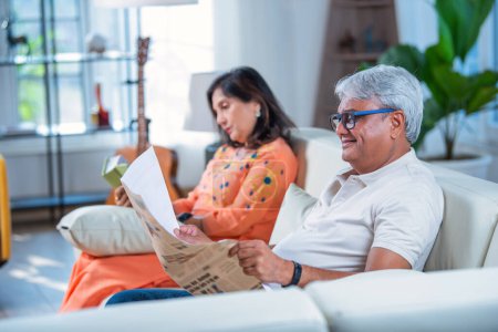 Téléchargez les photos : Personnes âgées indien asiatique couple lecture journal et livre tandis que assis sur canapé ou canapé dans salon - en image libre de droit