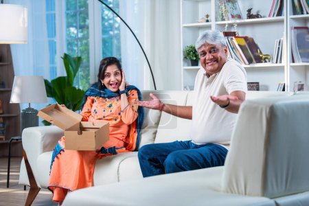 Téléchargez les photos : Senior indienne asiatique femme avec son mari ouvrir une boîte-cadeau tout en étant assis à la maison sur le canapé - en image libre de droit