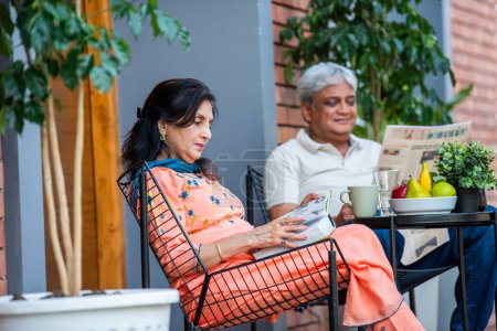 Téléchargez les photos : Personnes âgées indien asiatique couple lecture journal et livre tout en listning à la musique et avoir petit déjeuner à l'extérieur dans galerie - en image libre de droit