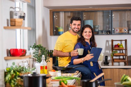 Téléchargez les photos : Indien heureux jeune couple en utilisant tablette PC dans la cuisine et prendre le thé ou le café tout en se tenant près de la table - en image libre de droit