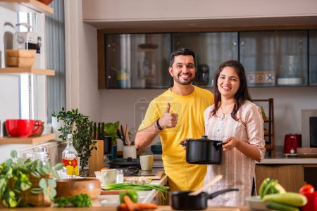 Téléchargez les photos : Heureux amour indien asiatique jeune couple tenant un pot et regardant caméra tout en cuisinant la nourriture dans la cuisine ensemble - en image libre de droit