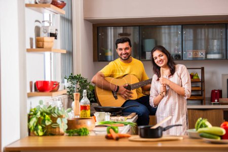 Photo for Indian young couple cooking food in kitchen, husband plays guitar and wife sings - Royalty Free Image