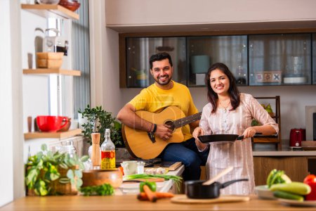 Foto de India pareja joven cocina comida en la cocina, marido toca la guitarra y esposa canta - Imagen libre de derechos