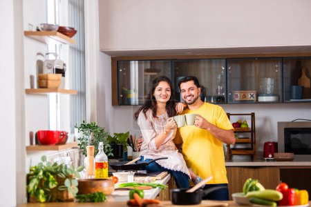Foto de India hermosa pareja joven tomando café en la cocina mientras cocina alimentos - Imagen libre de derechos