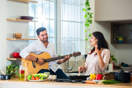 Photo for Indian young couple cooking food in kitchen, husband plays guitar and wife sings - Royalty Free Image