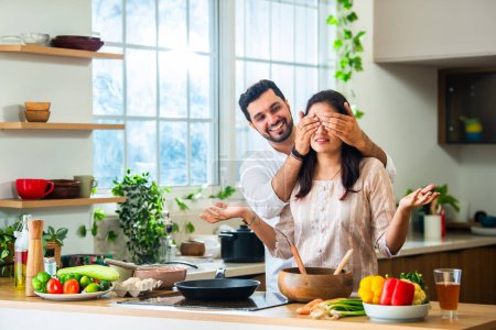 Téléchargez les photos : Indien jeune homme couvre ses yeux de femme faisant une surprise tandis que la femme occupé cuisine la nourriture dans la cuisine - en image libre de droit