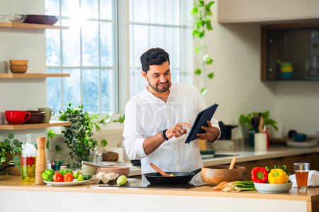 Téléchargez les photos : Indien beau mâle cuisine des aliments dans la cuisine et en utilisant tablette PC - en image libre de droit