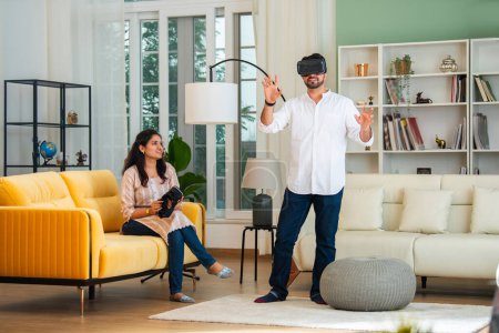Téléchargez les photos : Beau indien asiatique couple jouer jeu vidéo à l'intérieur à l'aide futuriste vr lunettes dans confortable maison cadre - en image libre de droit
